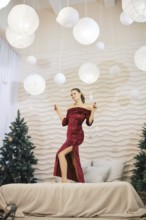 A stylish woman in a red dress stands playfully on a bed, surrounded by soft lights and Christmas
