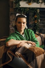 A woman wears a green sweater and a white ribbon in her hair while seated comfortably on a couch.