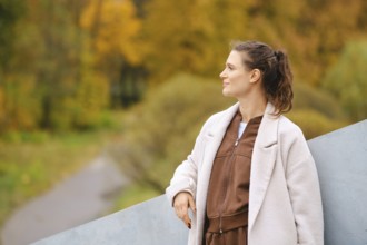 A woman stands calmly in an autumn landscape, surrounded by golden trees. She wears a cozy outfit