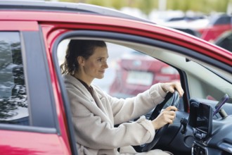 A woman sits in the driver's seat of a red car in a busy parking lot. She has a confident