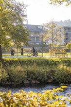 A cyclist rides through a park next to a river on a sunny autumn day, Urschelherbst Street