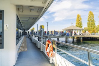 Harbour with people on a sunny day, boats in water, trees in shades of yellow and orange,