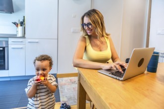 Woman working remotely on a laptop in her kitchen while taking care of her baby son eating an apple