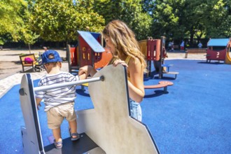 Young mother assisting her little son while he is carefully climbing on a playground structure,