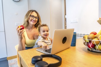 Mother holding an apple while son focused on laptop in kitchen, surrounded by a fruit basket,