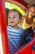 Smiling toddler playing joyfully on a playground slide while mother watches nearby, creating