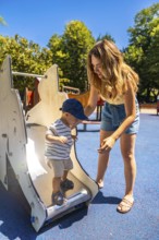 Mom carefully assisting her little boy while he's learning to use the slide at a children's