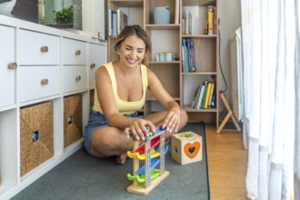 Young woman playing with a toy car ramp and smiling while sitting on a play mat in a child's