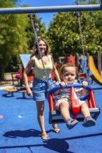 Young child enjoying a swing at the playground with their mother looking on, having fun on a summer