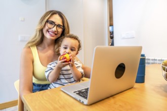 Mother holding her son while enjoying a nectarine and working on a laptop in the bright kitchen,
