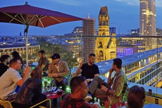 People on an outdoor terrace in the evening with a view of the illuminated Kaiser Wilhelm Memorial