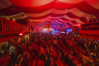 Full beer tent with people, red ceiling and stage, lively and festive atmosphere, Volksfest