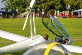 A motorised glider parked at the edge of the airfield with the canopy open during an air show at