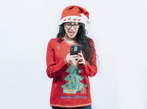 Amazed girl in Christmas hat looking at phone isolated. Surprised girl in Christmas sweater looking