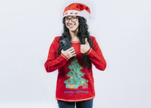 Smiling girl in Christmas hat holding phone with thumb up isolated. Positive young woman in