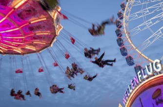 An illuminated chain carousel turns at night at a funfair, with floating people and colourful
