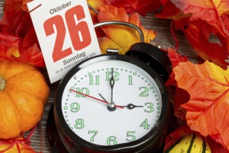 Time changeover to winter time 2025: Close-up of an alarm clock, autumnal decoration and a page