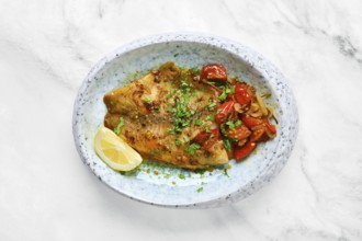 A perfectly cooked white fish fillet is topped with sautéed tomatoes and fresh herbs. A lemon wedge