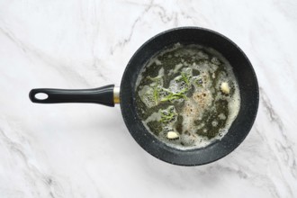 A frying pan is on a marble surface, filled with bubbling butter, fresh garlic and frosemary.