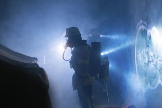 Firefighters in protective suits work in the fog, beams of light cut through the smoke,