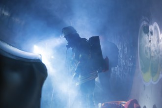 Firefighters with breathing apparatus shrouded in fog, illuminated by bluish light, firefighting