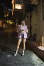 A woman stands on a cobblestone street at night, dressed in a pink blazer and casual shorts. Soft