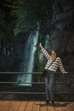 A cheerful young woman stands on a wooden walkway behind a railing, posing joyfully with one arm