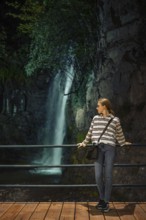 A young woman enjoys a quiet moment by a waterfall in a lush, natural setting during the evening