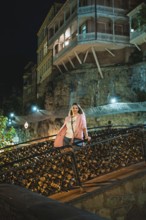A woman stands on a decorative bridge at night. The backdrop showcases illuminated historic