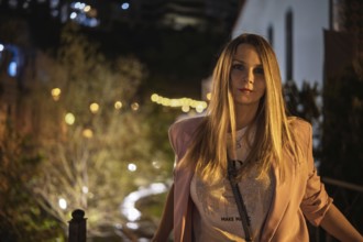 A woman poses on a pathway at night, dressed in a fashionable outfit with long hair. The scene