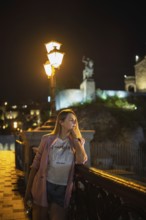 A woman stands by a decorative railing of the bridge, admiring the night scenery. Dim lights