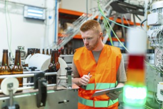Young man in safety vest checking beer bottles on a factory conveyor belt, performing quality