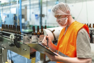 Worker in a hairnet and face mask checking clipboard records while brown beer bottles move along a
