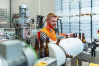 Smiling man in safety vest stands beside a conveyor belt, carefully inspecting brown glass bottles