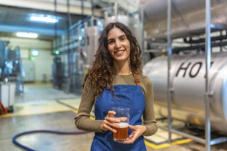 Woman brewer standing in modern brewery, smiling and holding a glass of craft beer for quality