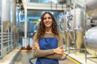 Woman brewer stands confidently with arms crossed, smiling at camera in a modern microbrewery with