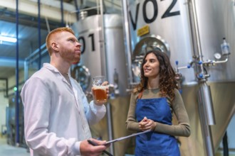 Two brewery professionals inspect and taste craft beer beside large fermentation tanks in a modern