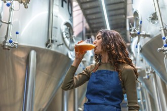 Woman wearing an apron tasting a glass of freshly brewed beer for quality control, standing in a