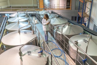 Woman scientist in a lab coat walking on a platform among large industrial fermentation tanks,