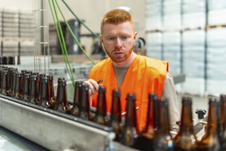 Man wearing an orange safety vest inspecting empty glass beer bottles moving along a conveyor belt