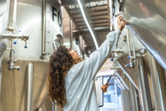 Young woman in a lab coat checking control panel on a stainless steel fermentation tank, performing