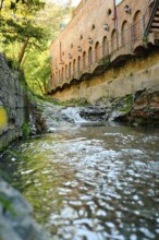 A clear stream flows calmly past old brick structures surrounded by lush greenery. Sunlight