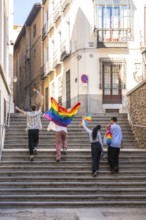 Young adults are joyfully walking up stone stairs in a european city, carrying rainbow flags and a