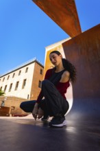 Young woman squatting on an urban surface, posing casually against a modern rust colored