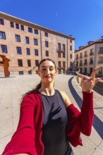 Young woman with dark hair posing for a selfie in a city square, holding her hand up, with old