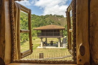 Open wooden window with rope hinges framing a scenic view of a traditional elevated horreo granary