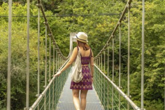 Female tourist with hat and backpack walking on a metal suspension bridge, enjoying the scenic