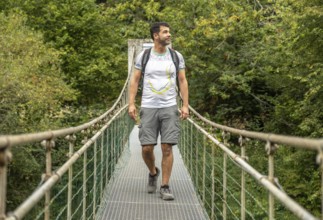 Male hiker with backpack enjoying trekking in the forest, walking on a metallic hanging bridge over
