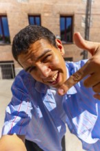 Young man smiling into camera and pointing down in a bright outdoor street selfie, casual striped