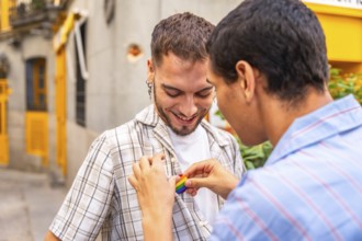 Young gay man gently attaching a rainbow pride flag pin to the plaid shirt of his smiling partner,
